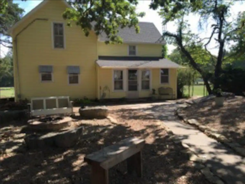 A yellow two-story house with a stone pathway and benches in a grassy area surrounded by trees.