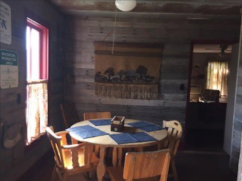 A rustic dining area with a round table, wooden chairs, and a woven wall hanging in a cozy, cabin-like setting.