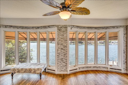 Bright room with large bay windows overlooking a lake, featuring a ceiling fan and wooden flooring.