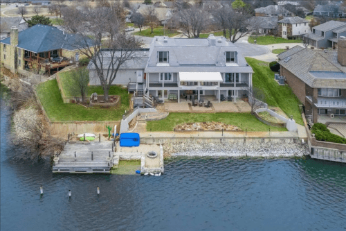 Aerial view of a waterfront home with a green lawn, patio, and dock, surrounded by other houses and trees.