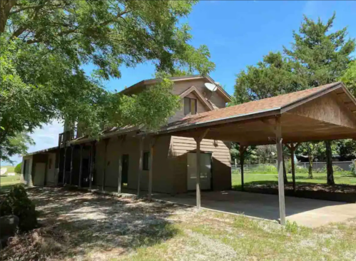 A two-story wooden house with a covered porch, surrounded by trees and a grassy area.