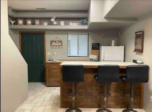 A cozy kitchen with a bar, three stools, and beach-themed decor on the walls. A green door is visible in the background.