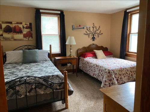 A cozy bedroom with two beds, a nightstand, and decorative wall art, featuring natural light from two windows.