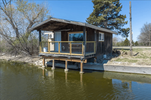 A small wooden cabin on stilts by a calm body of water, surrounded by trees and grass under a clear blue sky.