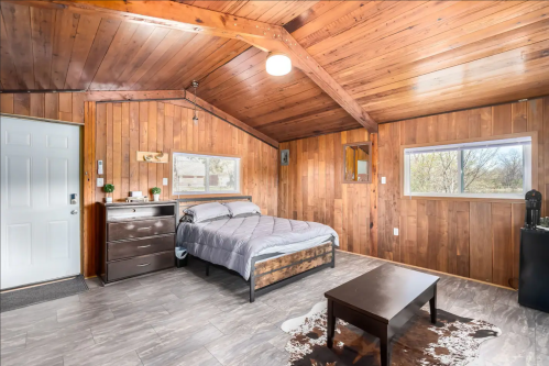 Cozy wooden bedroom with a bed, dresser, and large windows, featuring a rustic design and natural light.