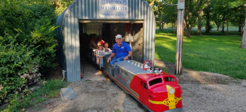 A small red train exits a shed, carrying children and an adult, surrounded by greenery in a park setting.