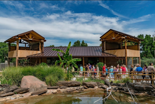 A crowd gathers outside a modern building with wooden features and large windows, surrounded by greenery and a pond.
