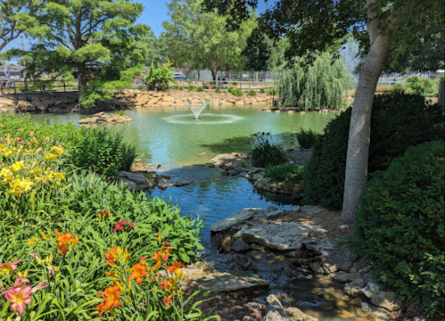 A serene pond surrounded by vibrant flowers and lush greenery, with a fountain in the center and trees in the background.