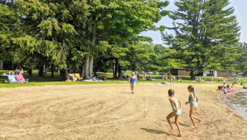 Two children run on a sandy beach while adults relax under trees in the background. Bright, sunny day.