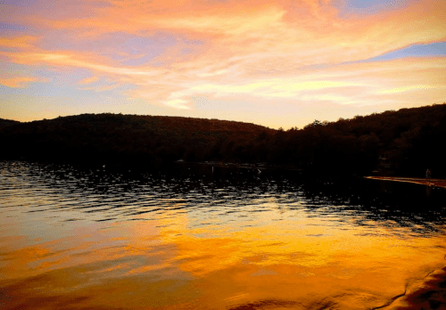 A serene lake at sunset, reflecting vibrant orange and purple hues with hills in the background.