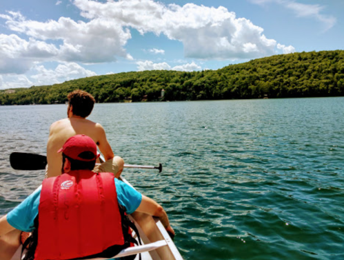 Two people in a canoe on a lake, surrounded by green hills and a blue sky with fluffy clouds.