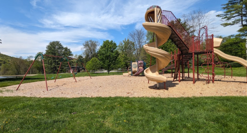 A playground featuring a spiral slide, swings, and climbing structures, surrounded by green grass and trees.