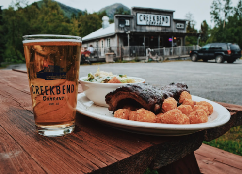 A glass of beer next to a plate of ribs and tater tots, with a rustic restaurant in the background.
