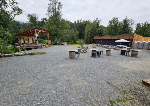 Outdoor event space with gravel ground, wooden stage, tables, and trees in the background. Restrooms visible on the side.