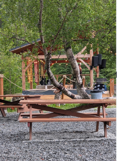 A wooden picnic table in a gravel area, with a gazebo and trees in the background, suggesting an outdoor gathering space.