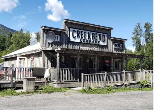 A rustic wooden building with a sign reading "Creek Bend Company," featuring a porch and surrounded by greenery.