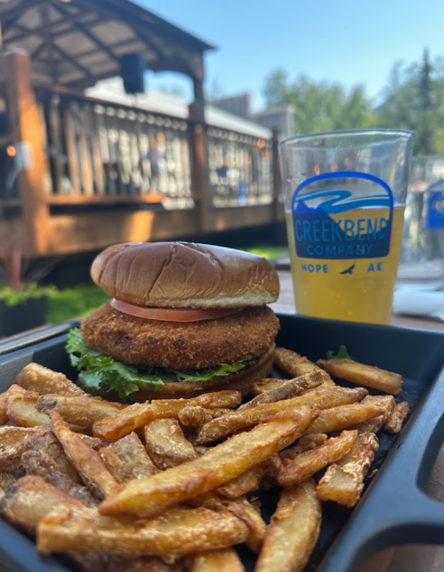 A crispy chicken sandwich with lettuce and tomato, served with golden fries and a glass of beer on a sunny patio.