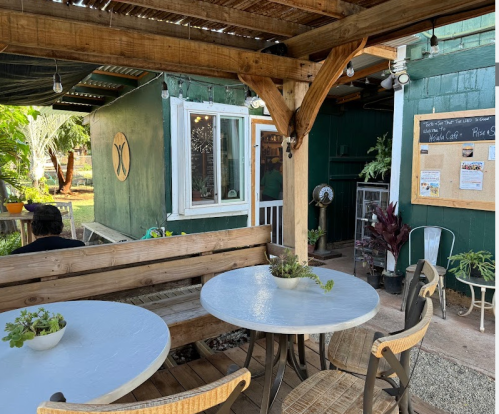 A cozy outdoor seating area with a white table, chairs, and plants, next to a green building and bulletin board.