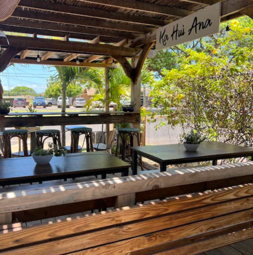 A cozy outdoor seating area with wooden benches and tables, surrounded by greenery and a sign that reads "Ka Hui Ana."