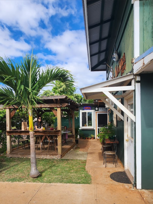 A cozy outdoor café with a palm tree, wooden seating, and a blue sky in the background.