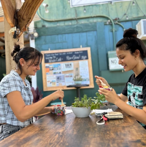 Two women sit at a wooden table enjoying ice cream, surrounded by plants and a chalkboard menu in a casual outdoor setting.