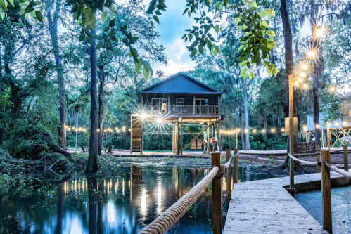 A wooden house on stilts beside a calm waterway, surrounded by trees and illuminated by string lights at dusk.