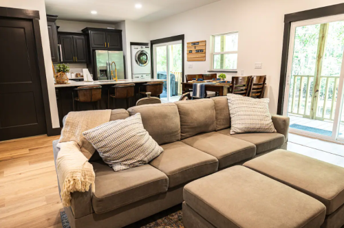 Cozy living room with a beige sectional sofa, decorative pillows, and a view of a kitchen and outdoor area.