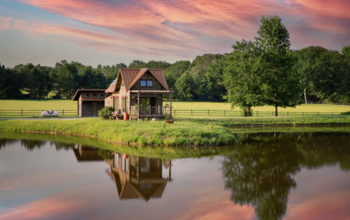 A quaint house by a pond, surrounded by lush greenery and a colorful sunset sky.