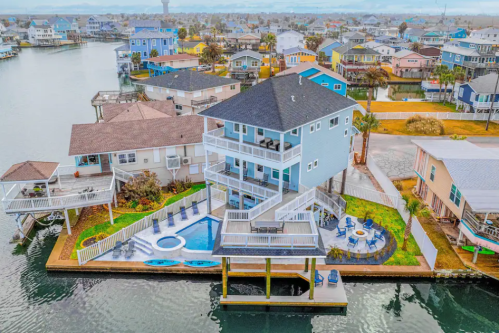 Aerial view of a modern waterfront house with a pool, surrounded by colorful homes and water.