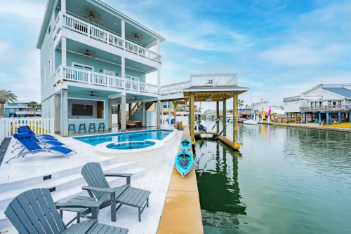A waterfront property with a pool, dock, and kayaks, featuring a spacious deck and blue sky in the background.