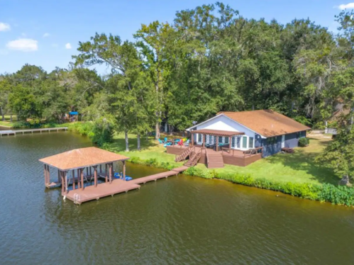 Aerial view of a lakeside house with a dock, surrounded by trees and a clear blue sky.