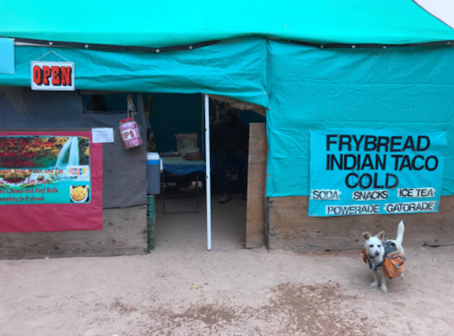 A food stand with a turquoise tent, featuring signs for frybread tacos, snacks, and drinks, with a dog in a backpack.