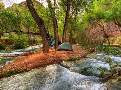 A serene campsite by a flowing river, featuring a tent and hammocks nestled among lush green trees.