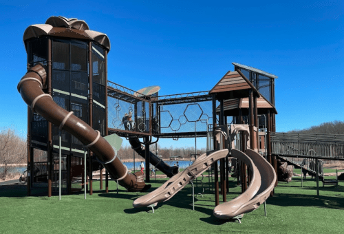 A modern playground with a large slide, climbing structures, and a grassy area, set against a clear blue sky.