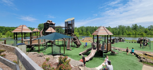 A vibrant playground with slides and climbing structures, surrounded by greenery and a lake under a clear blue sky.