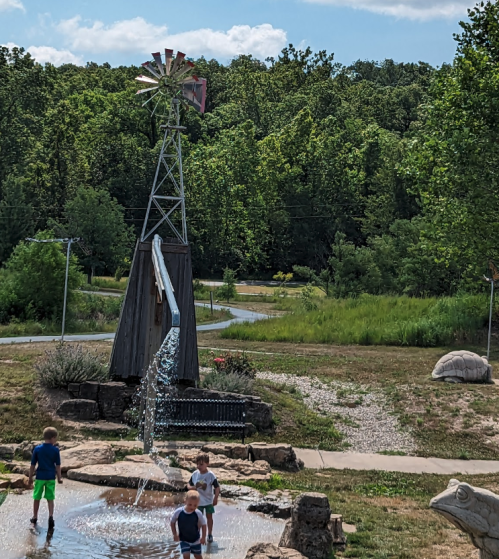 Children play in a water feature with a windmill in a lush green park under a blue sky with clouds.