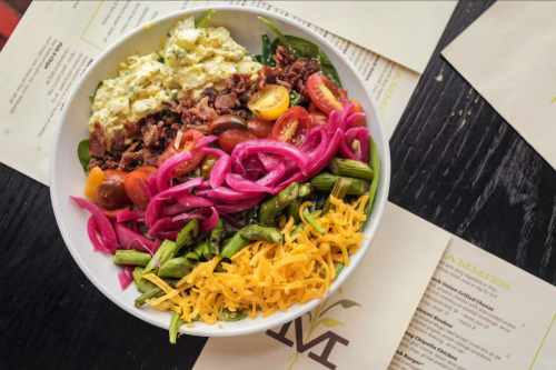 A colorful salad bowl with greens, tomatoes, pickled onions, cheese, and a creamy topping, surrounded by menus.