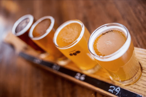 A wooden flight board holds four small glasses of different beers, showcasing a variety of colors and styles.