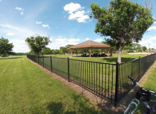 A fenced park area with a gazebo surrounded by green grass and trees under a blue sky with clouds.