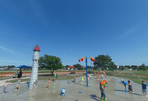 Children playing in a splash pad with water features under a clear blue sky. Trees and park benches in the background.