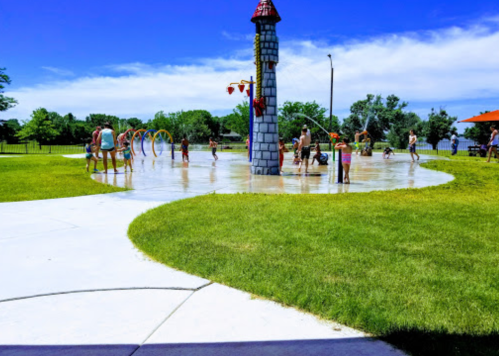 Children and adults play in a splash pad with colorful water features under a clear blue sky.
