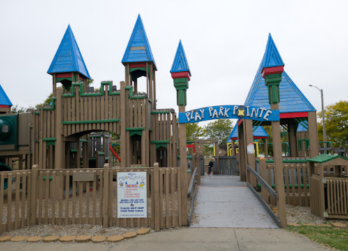 Colorful playground with blue-roofed structures and a sign reading "Play Park Pointe." Surrounded by wooden fencing.
