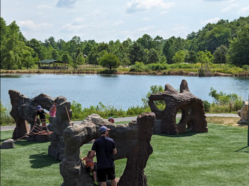 Children play on rock-like structures near a serene lake surrounded by greenery on a sunny day.