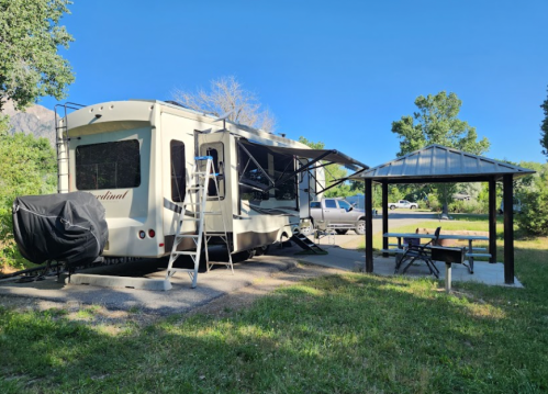 A parked RV with an awning, a picnic area, and a ladder nearby, surrounded by trees and clear blue skies.