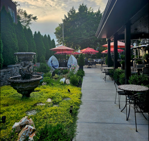 A scenic outdoor patio with a fountain, greenery, and red umbrellas over tables, set against a cloudy sky.