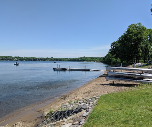 A serene lakeside view with a sandy shore, a dock, and boats on calm water under a clear blue sky.