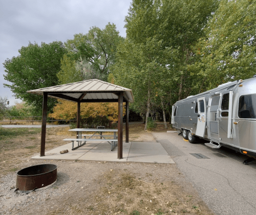A campsite with a picnic table under a gazebo, an Airstream trailer parked nearby, surrounded by trees.