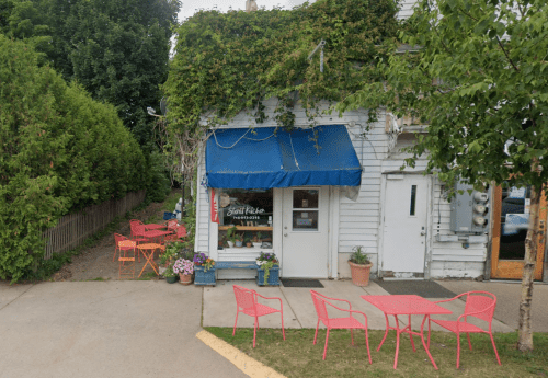 A small shop with a blue awning, surrounded by greenery, featuring outdoor seating with red tables and chairs.