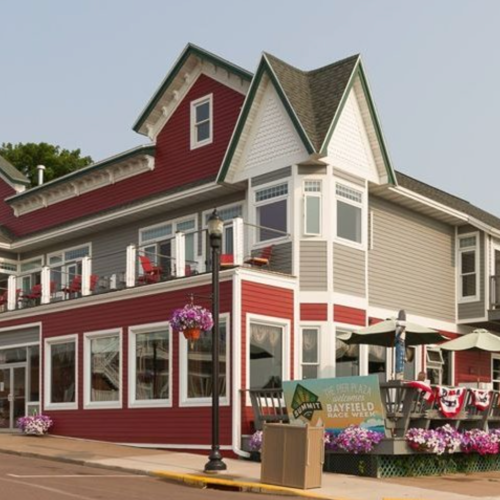 Charming red and gray restaurant with a balcony, flower planters, and outdoor seating on a sunny day.