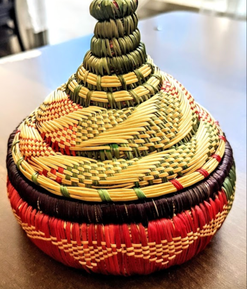 A colorful woven basket with a conical lid, featuring intricate patterns in green, red, and yellow.